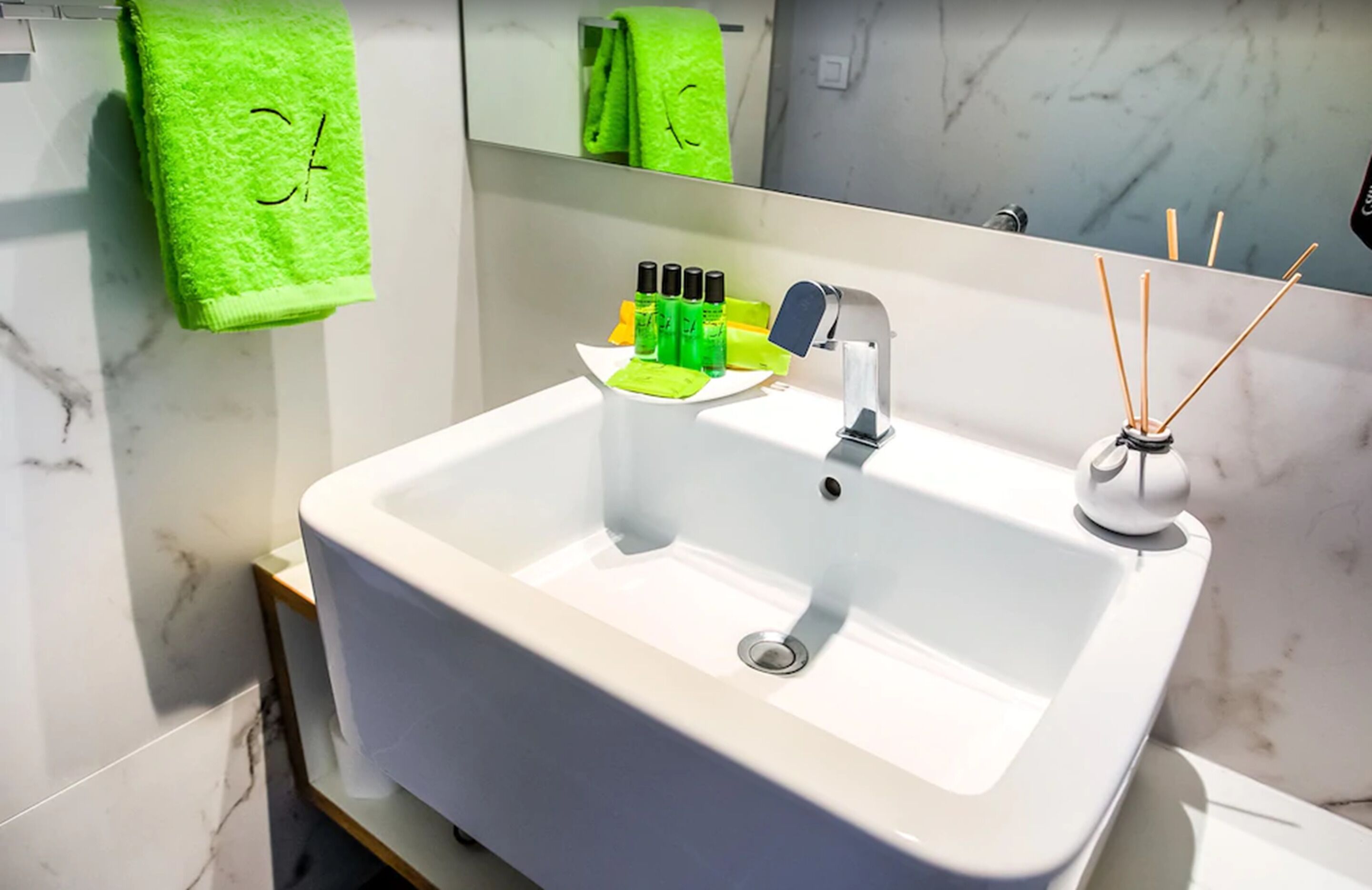 Un baño con un lavabo blanco, toallas verdes, un espejo y un difusor de aroma de caña.