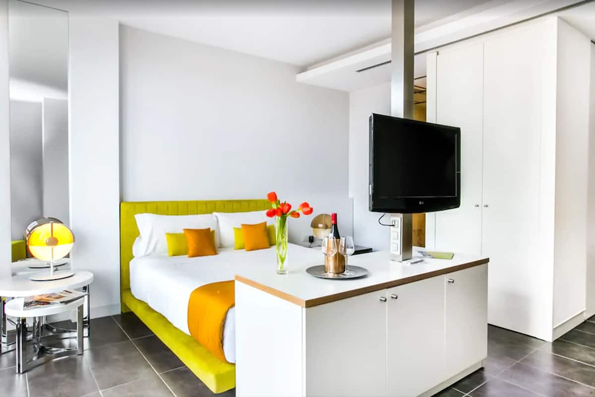 A modern hotel room with a bed, a TV, a small table with a lamp, and a minibar.