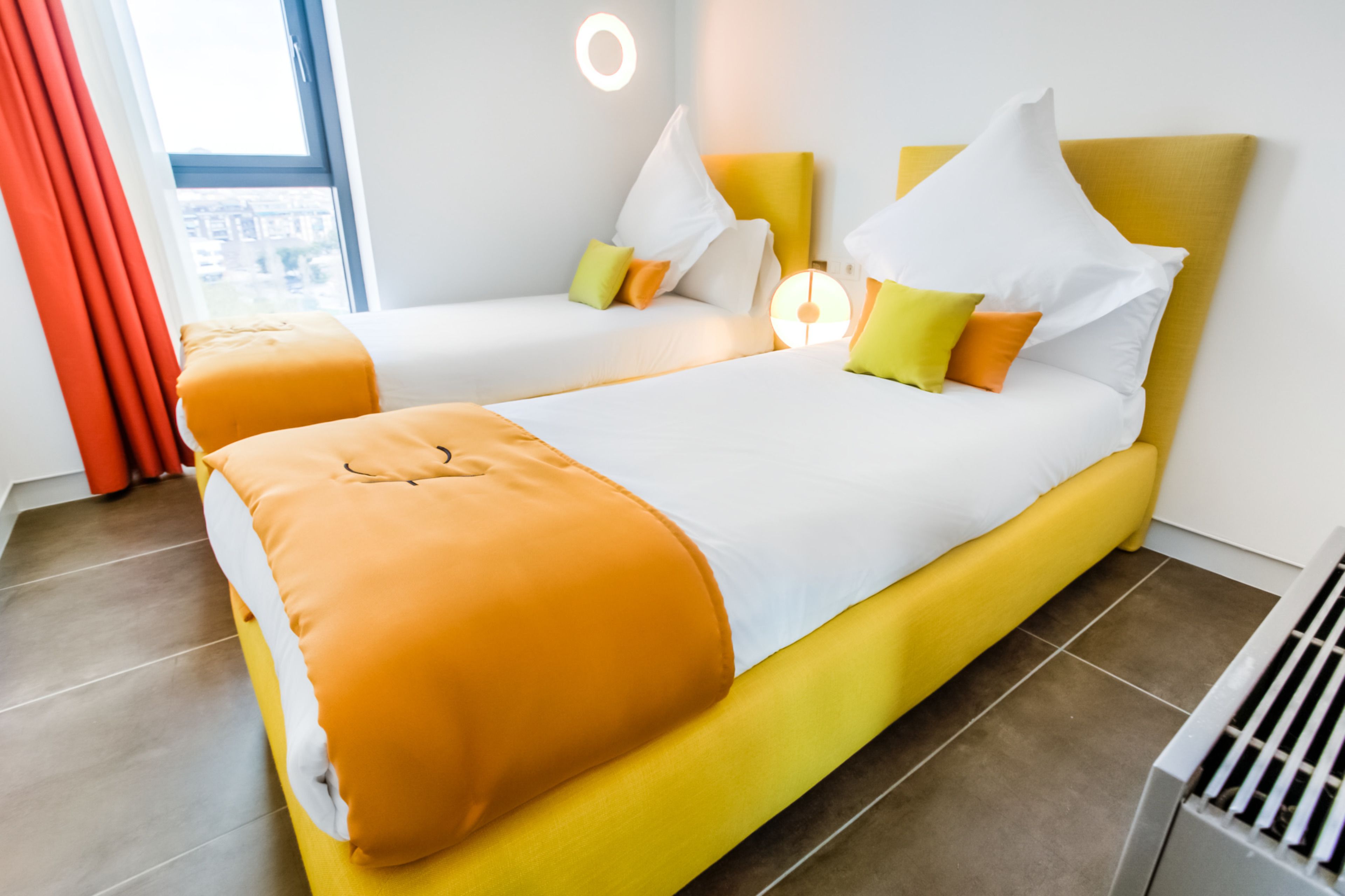 Ein Hotelzimmer mit zwei Betten, jeweils mit gelb-weißer Bettwäsche, und einem großen Fenster mit roten Vorhängen.