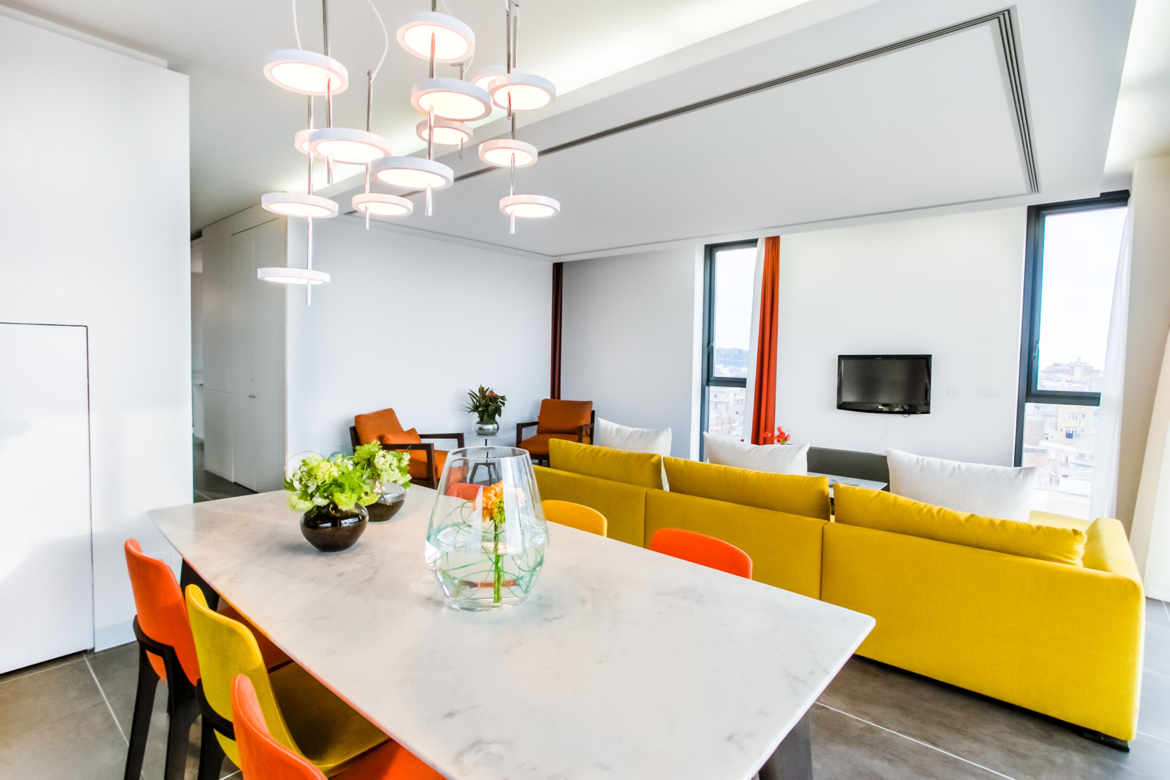 A modern living room with a yellow sofa, a dining table with chairs, and a large window with curtains.