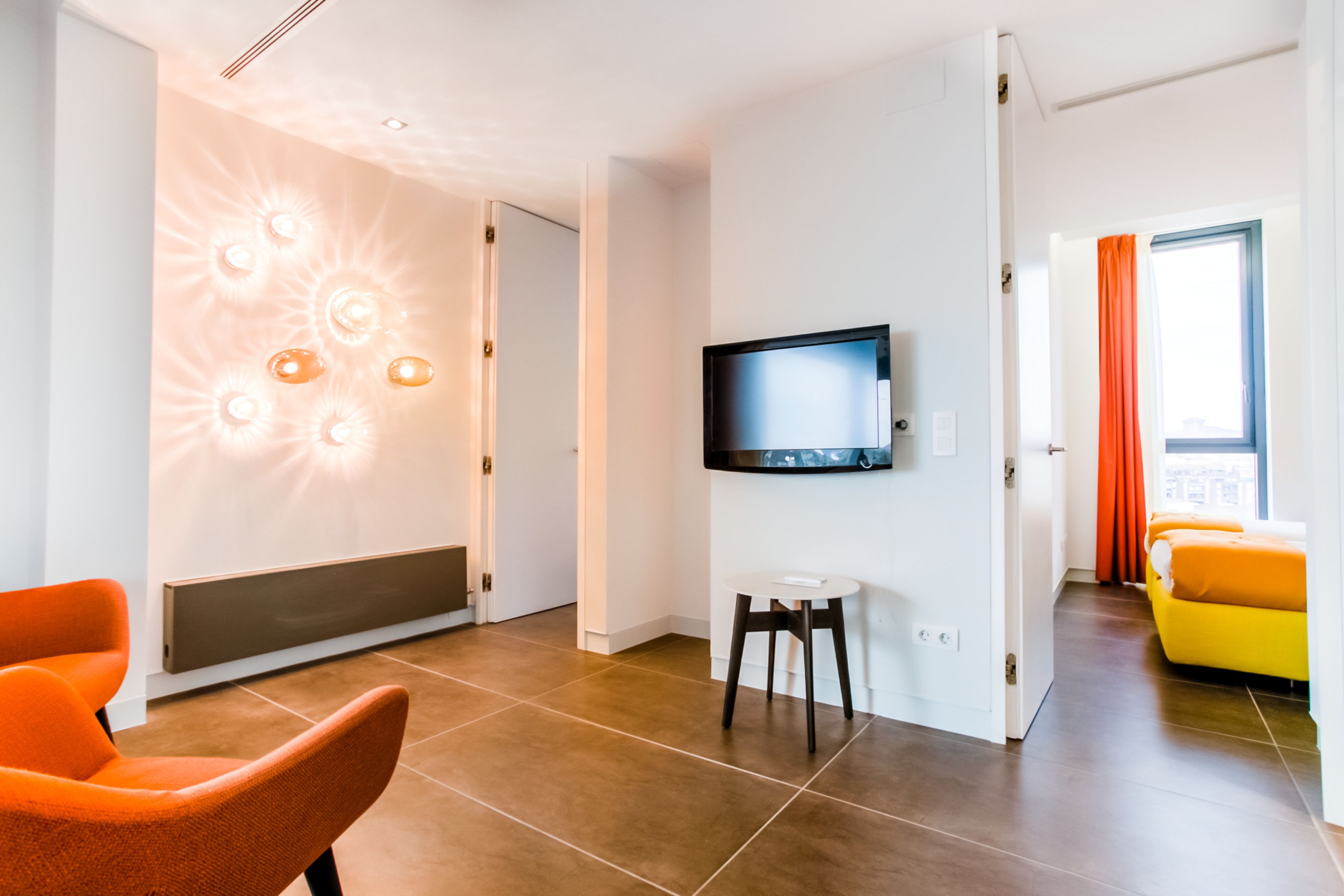 A modern hotel room with a flat-screen TV mounted on the wall, a seating area with a chair and a small table, and a bed with orange bedding.