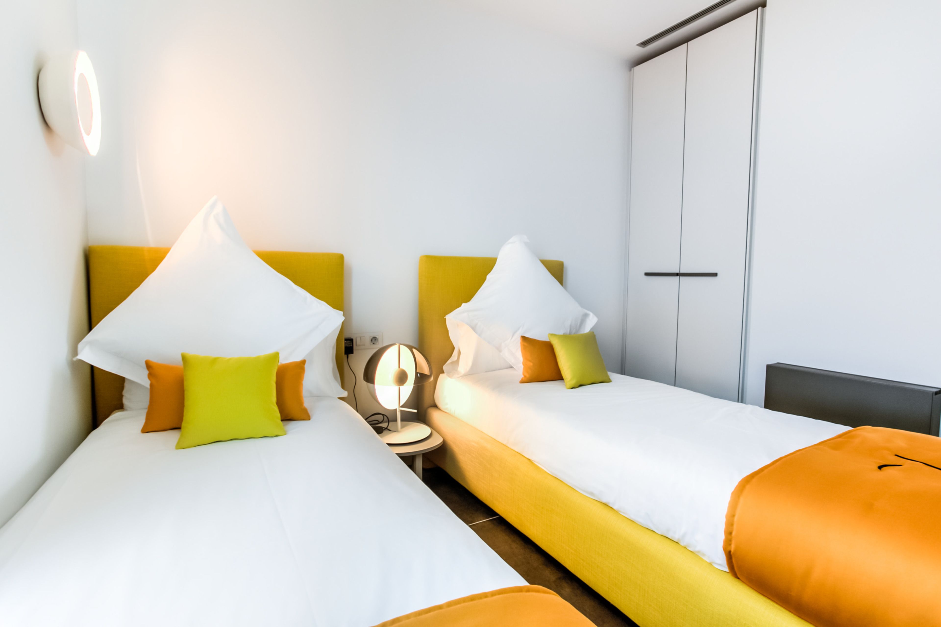 Quarto de hotel com duas camas, cada uma com roupa de cama branca e cabeceiras amarelas. Há uma mesa de cabeceira com uma luminária e uma mesinha redonda com uma cadeira.