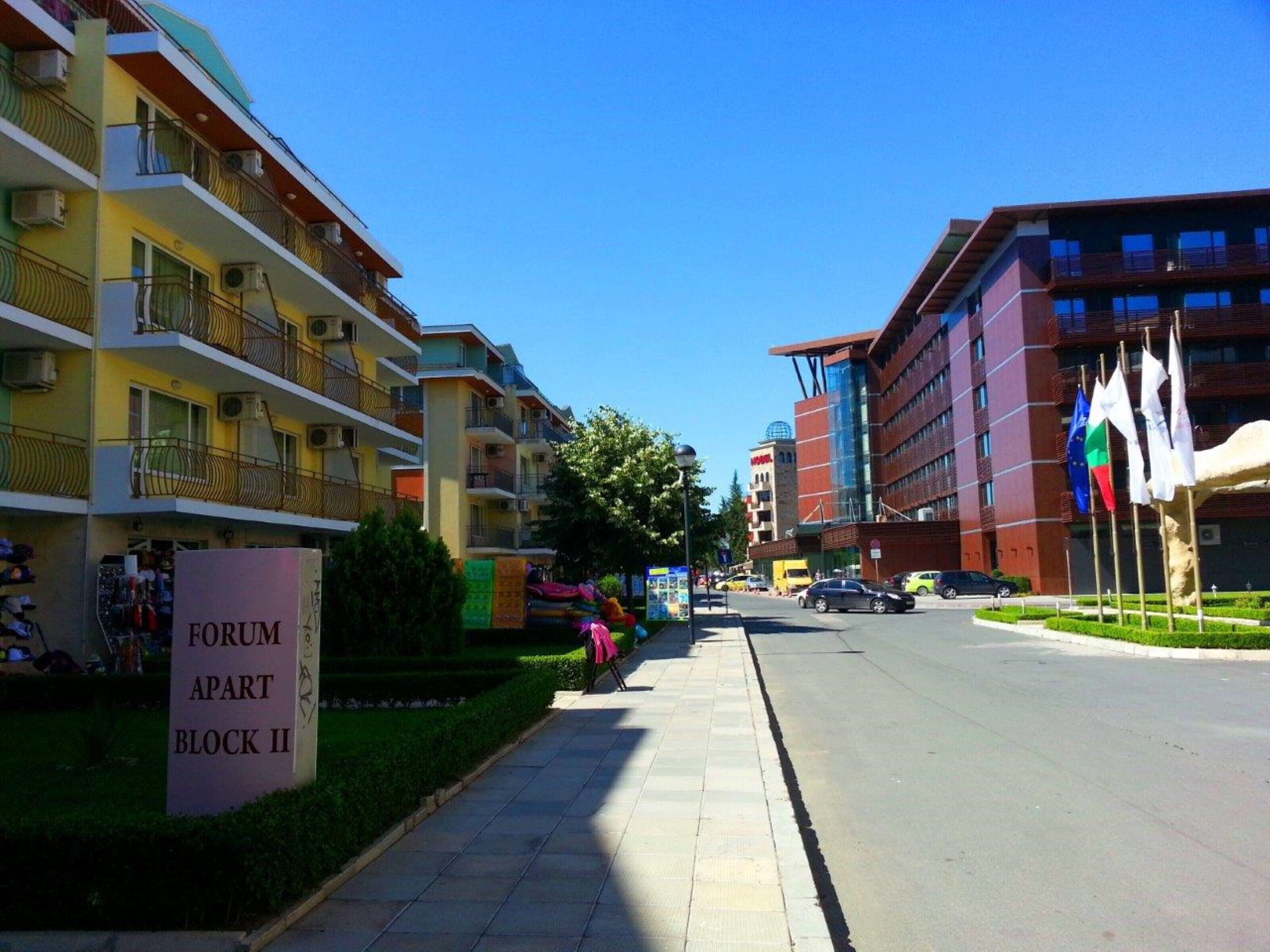 A multi-story residential building with balconies, a sign for "FORUM APART BLOCK II," and a street with cars and flags.