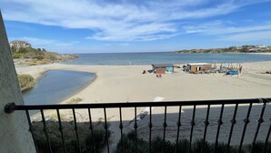 A beach view from a balcony with a railing, sandy beach, calm sea, and a few buildings in the background. - Menada Rocamar Apartments (Tsarevo)