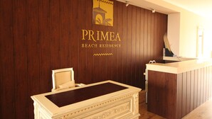 A reception desk with a wooden panel wall, a sign reading "PRIMEA BEACH RESIDENCE," and a small seating area with a chair and a table. - Menada Rocamar Apartments (Tsarevo)