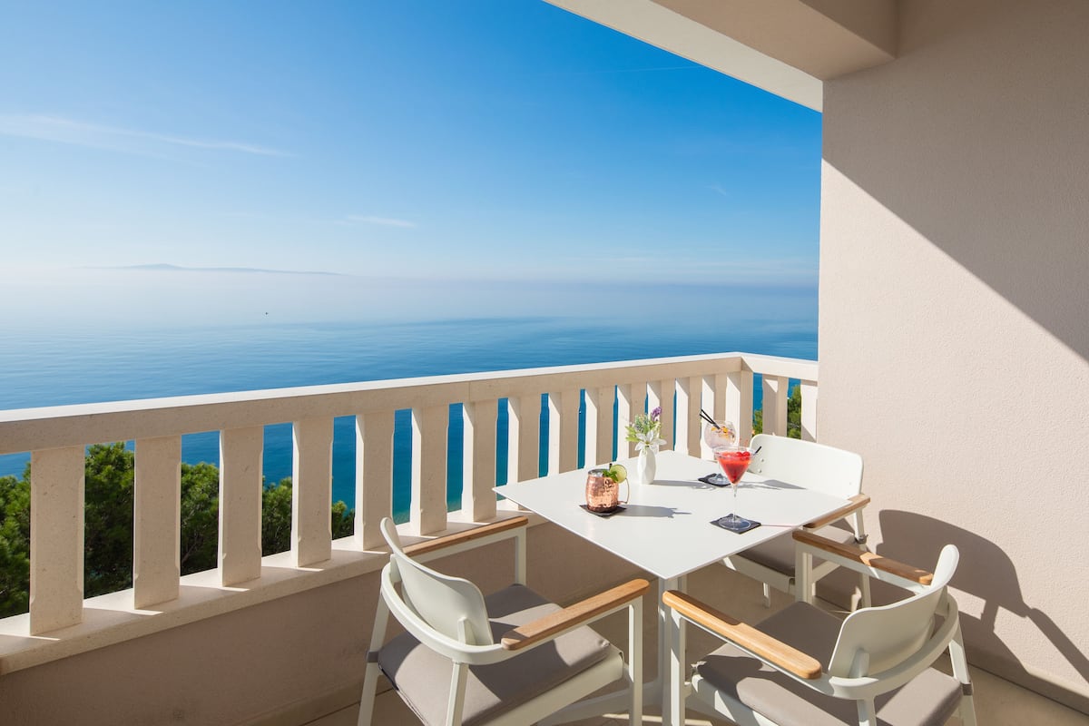 A balcony with a white table and chairs, overlooking a clear blue sea.