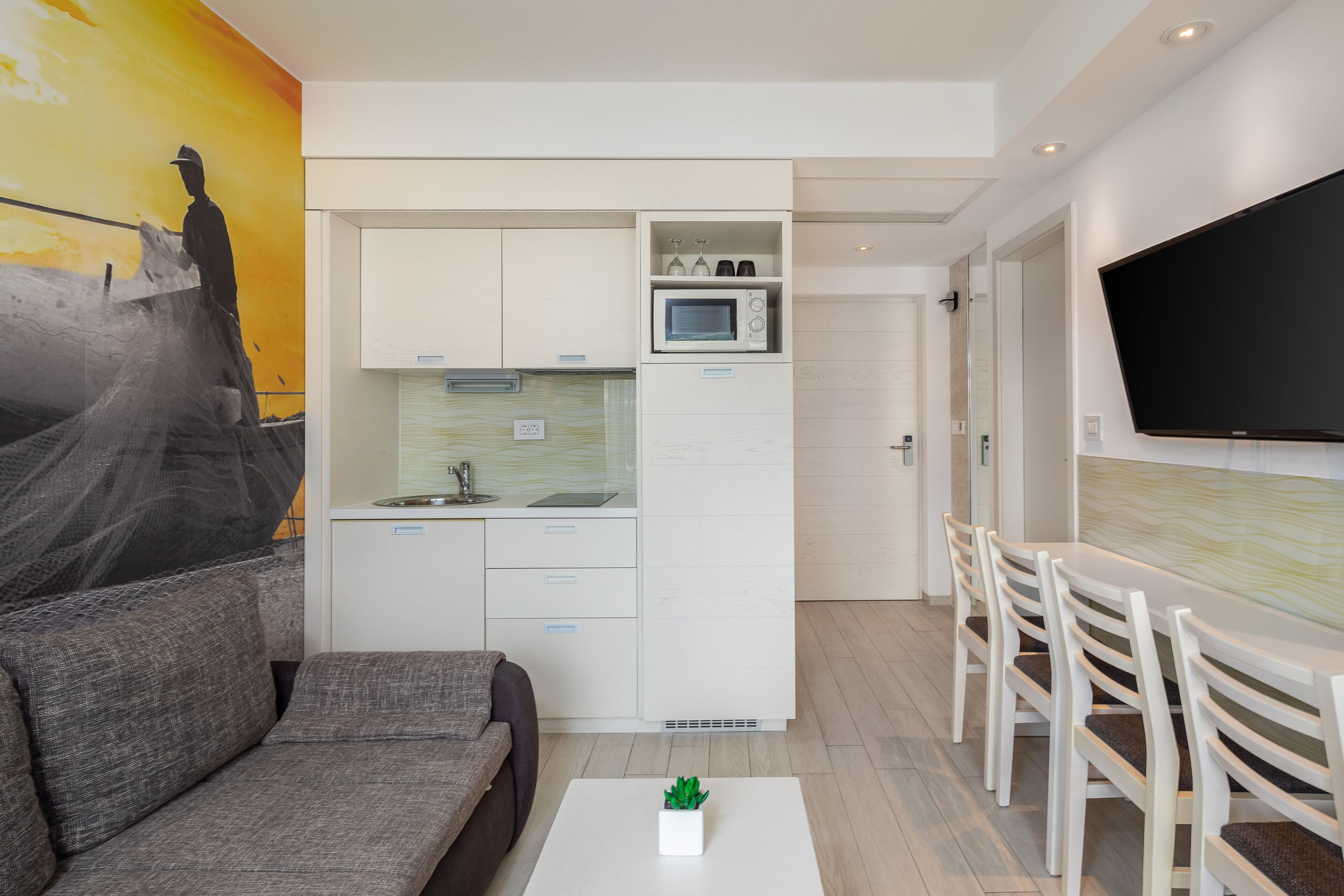 A compact living space with a kitchenette, a flat-screen TV mounted on the wall, a gray sofa, and a dining area with white chairs.