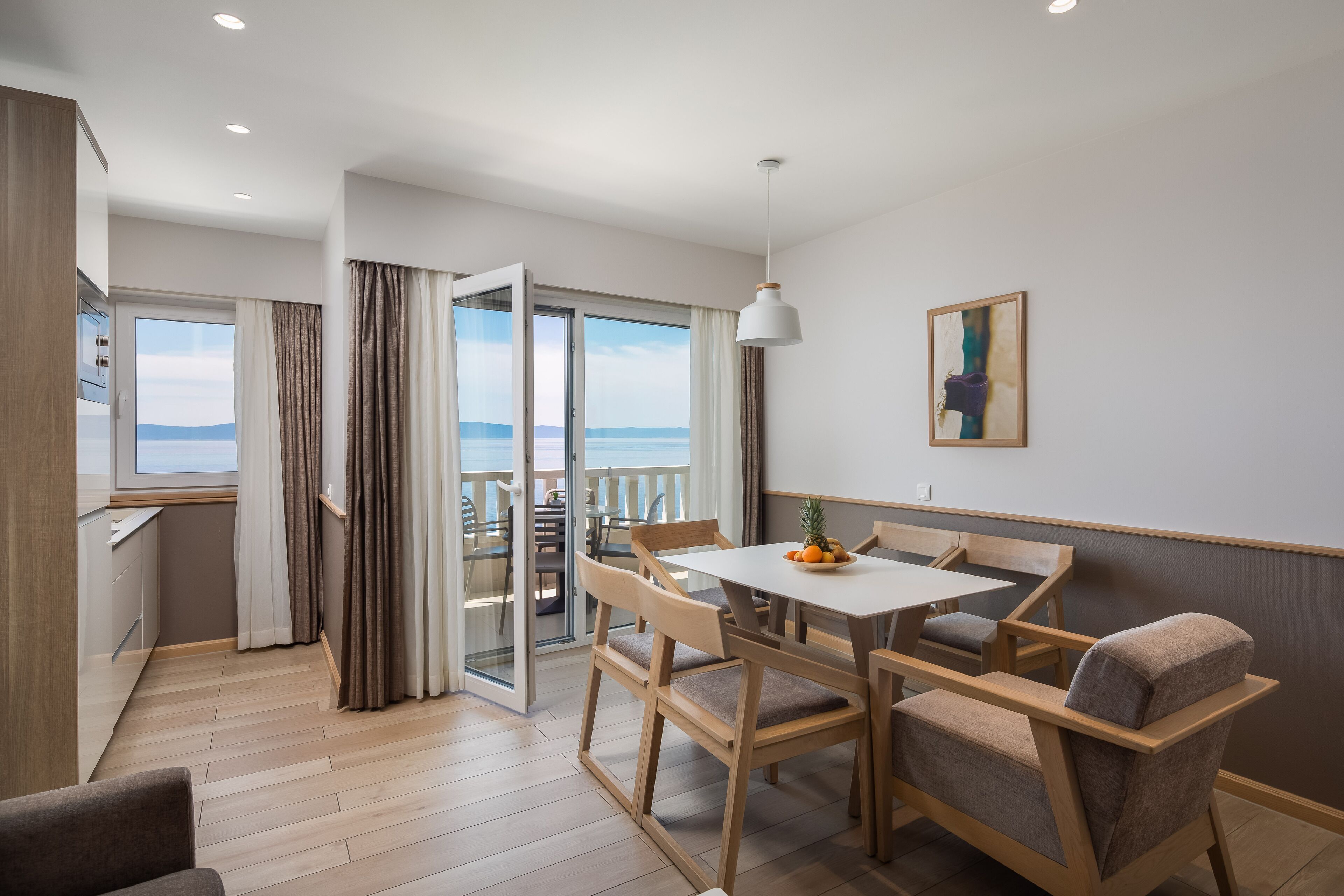 A modern dining area with a table and chairs, a view of the sea through large windows, and a balcony with a sliding door.