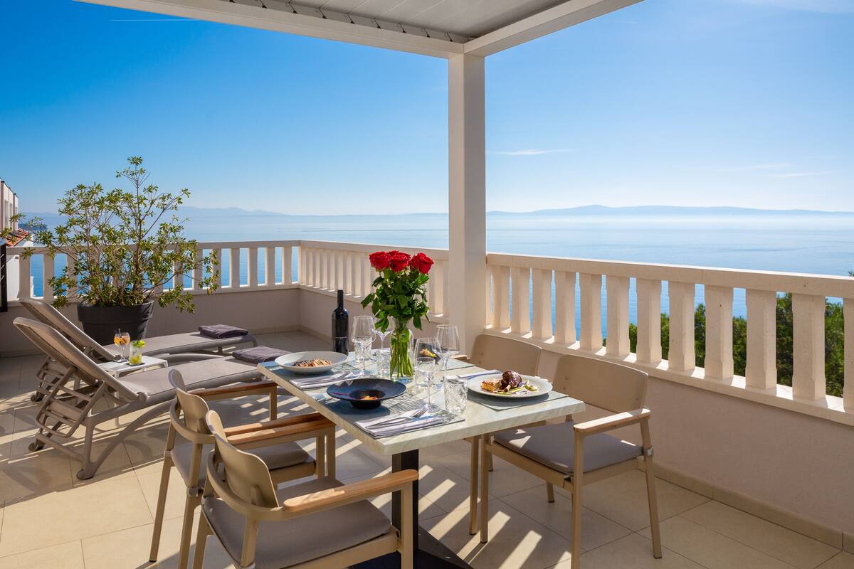 A balcony with a table set for dining, chairs, a potted plant, and a view of the sea.