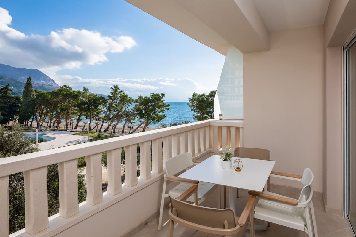 A balcony with a white table and chairs, overlooking a pool and trees with a clear view of the sea and mountains.
