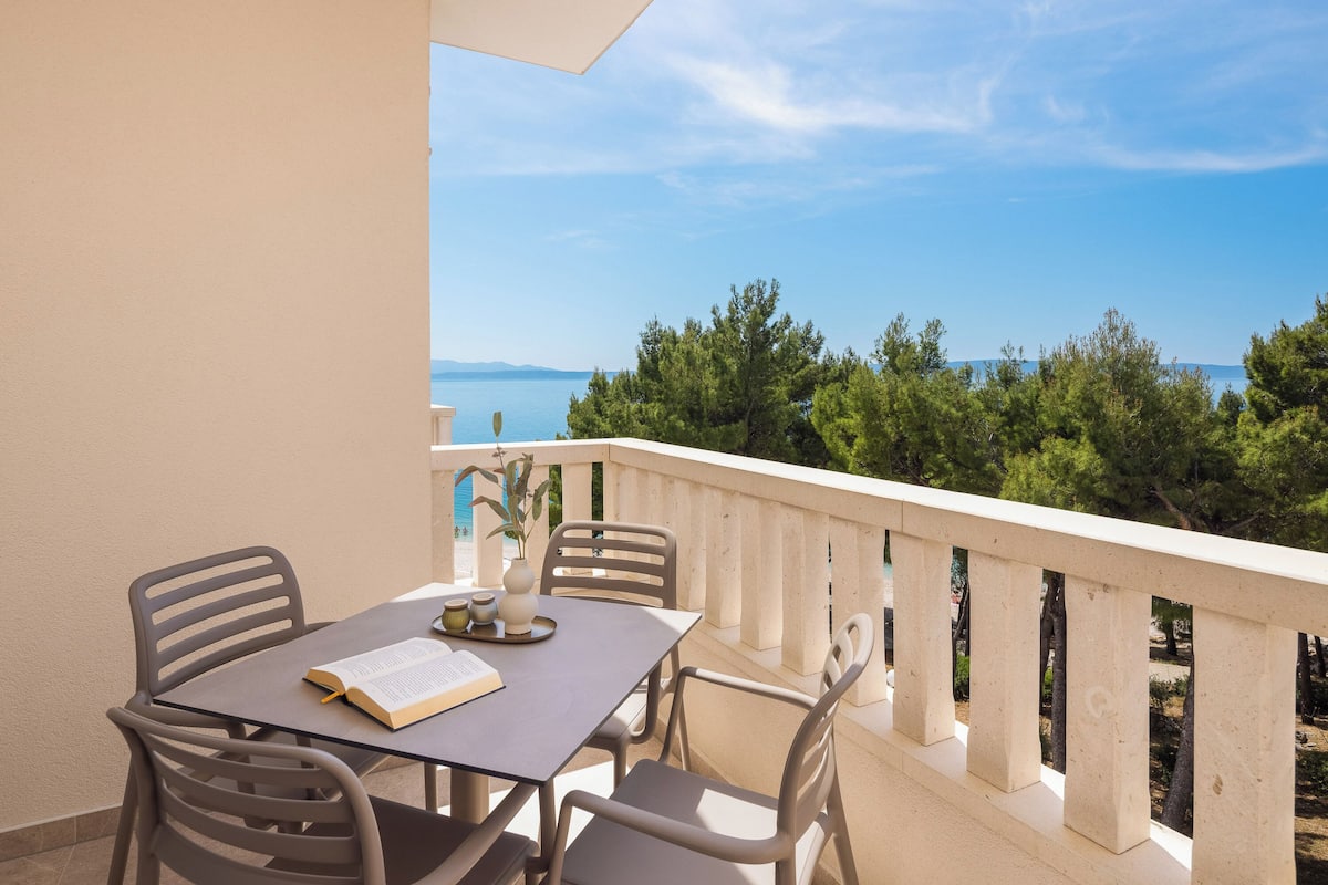 A balcony with a table and chairs, overlooking a coastal landscape with trees and the sea.