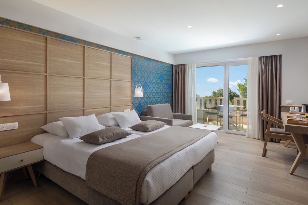 A hotel room with a large bed, a desk with a chair, a balcony with a table and chairs, and a view of the outdoors.