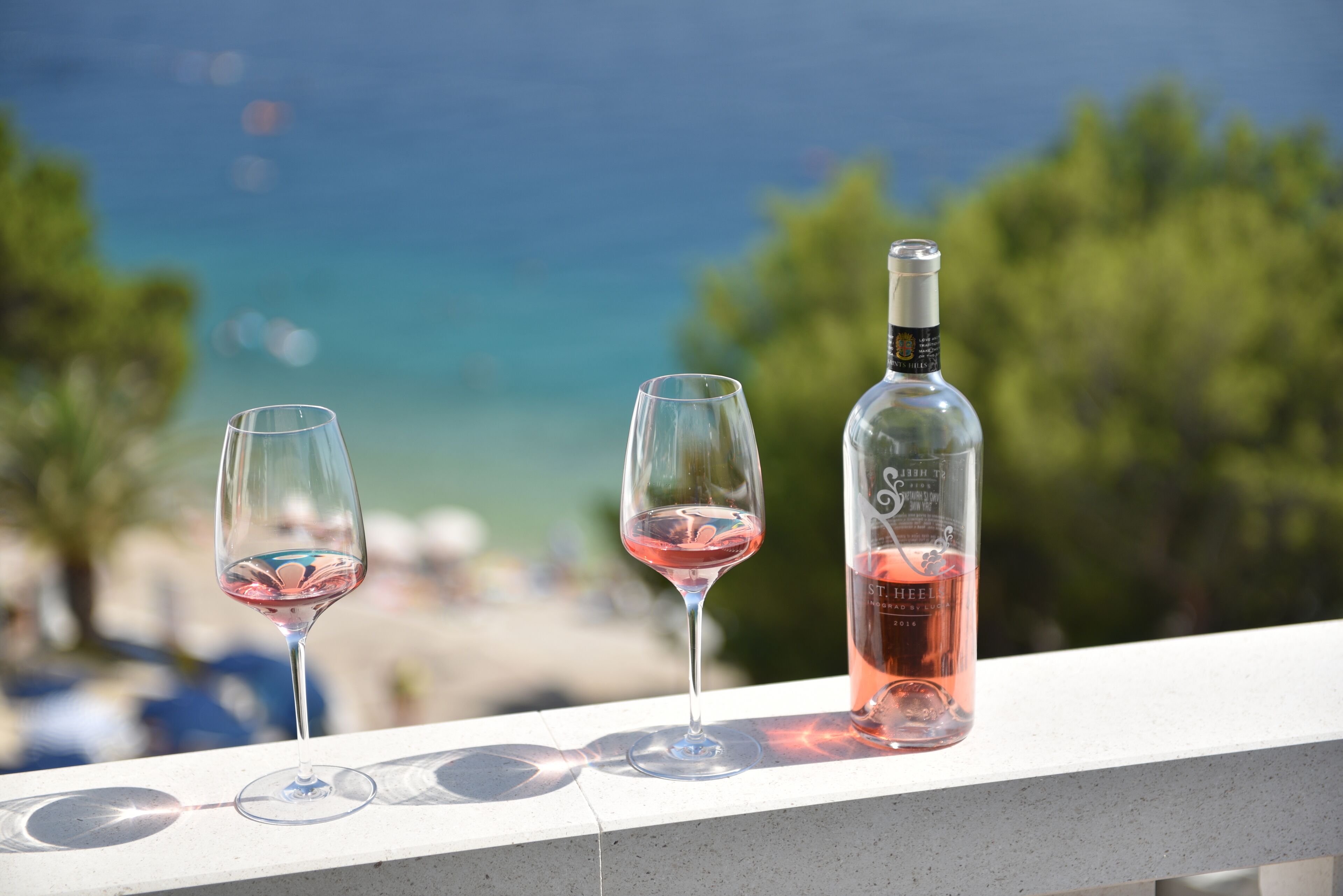 Three wine glasses and a bottle on a balcony with a view of the sea and trees.