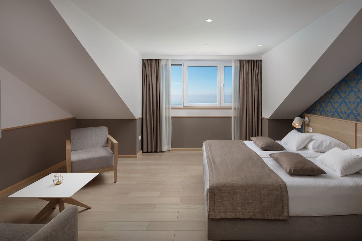 A modern hotel room with a bed, a chair, a small table, and a window with curtains.