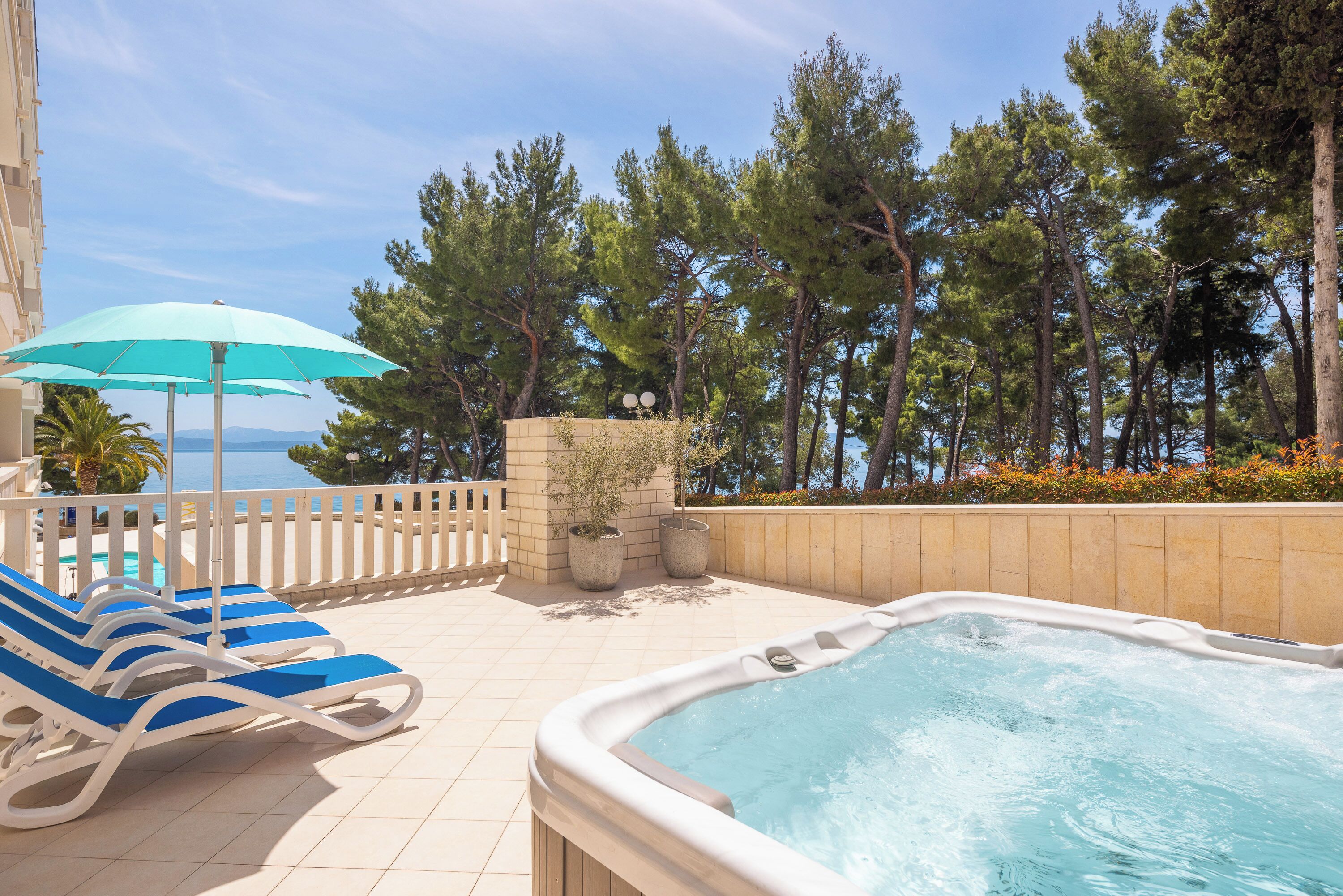 A pool area with lounge chairs and a hot tub, surrounded by trees and a view of the sea.