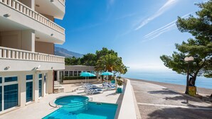 Area piscina con lettini e ombrelloni, vista libera sul mare e un edificio con balconi.