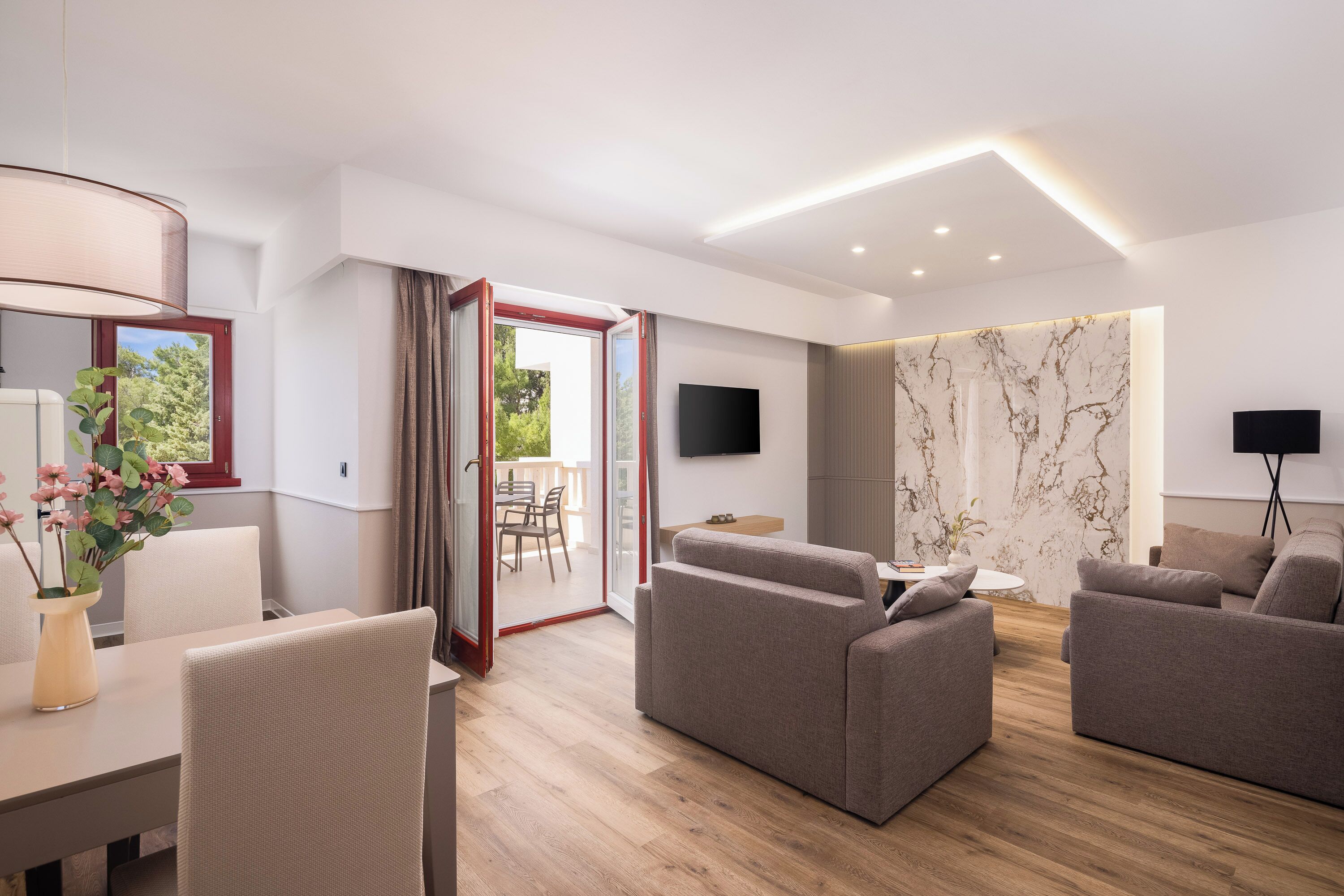 A modern living room with a sofa set, a dining table with chairs, a TV mounted on the wall, and a sliding glass door leading to an outdoor area.