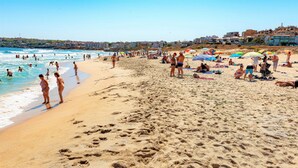 Ein belebter Strand mit badenden und sonnenden Personen, im Hintergrund sind Gebäude zu sehen.
