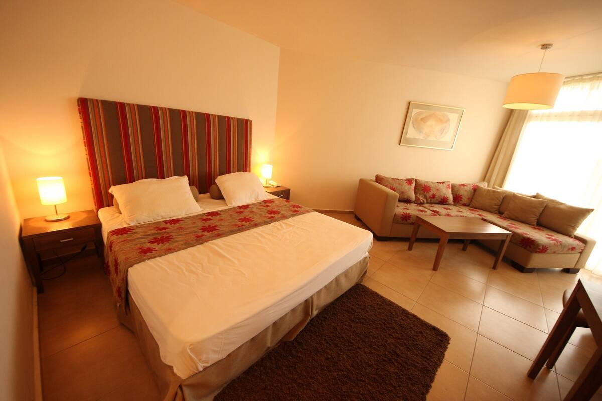 A hotel room with a bed, a sofa, a small table, and a nightstand with a lamp.