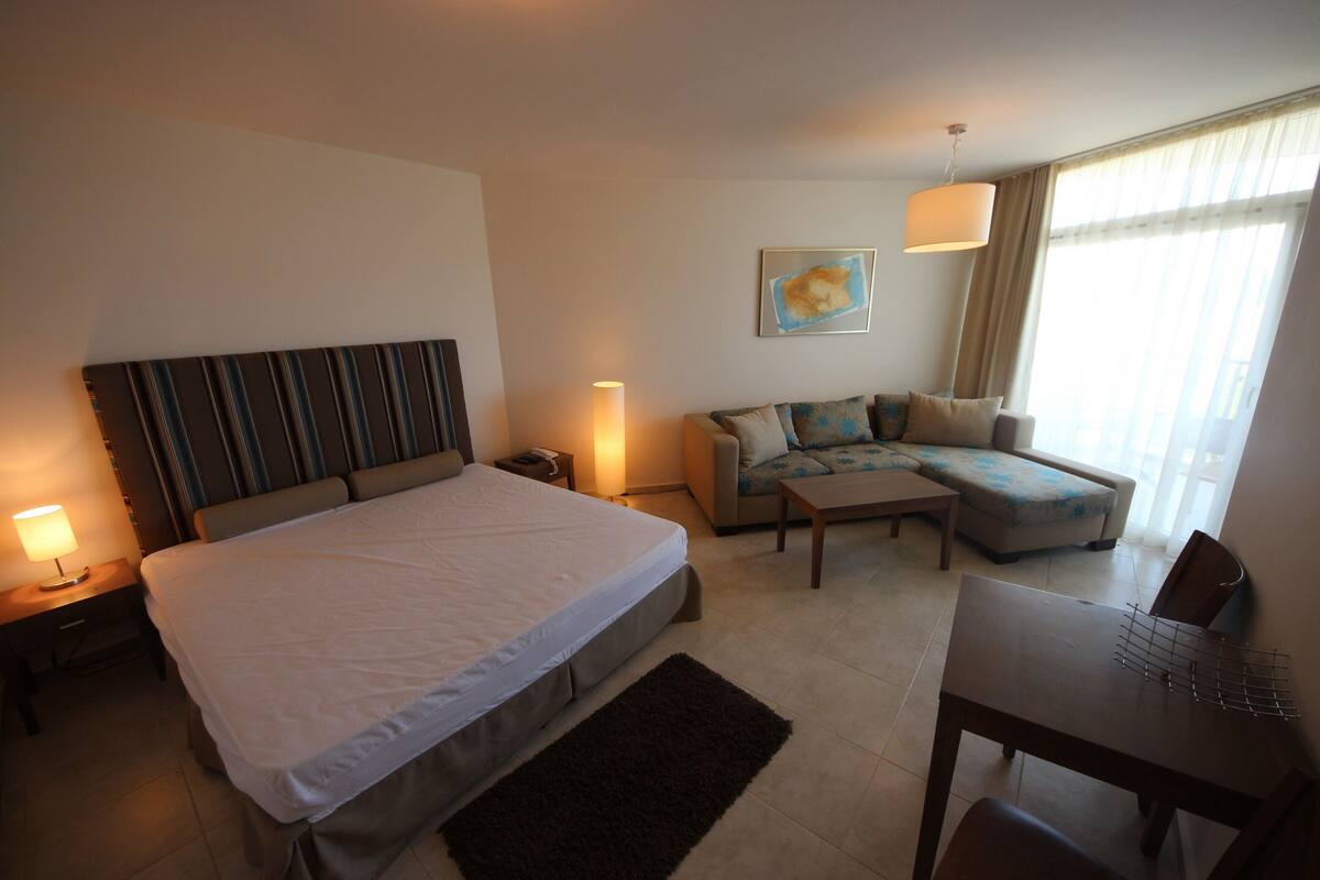 A hotel room with a large bed, a sofa, a dining table, and a lamp.