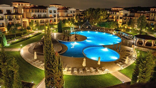 A large, illuminated swimming pool surrounded by lounge chairs and umbrellas, with a well-maintained lawn and buildings in the background.