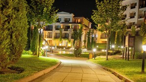 A nighttime view of a residential area with a paved pathway leading to a building with multiple balconies and a well-lit entrance.