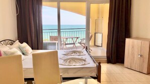 A hotel room with a bed, a glass table, a view of the ocean, a balcony with a table and chairs, and a wardrobe. - Menada Grand Resort Apartments (Pomorie)