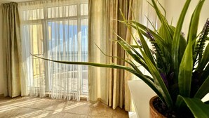 A room with a large window, curtains, a potted plant, and a chair. - Menada Grand Resort Apartments (Pomorie)