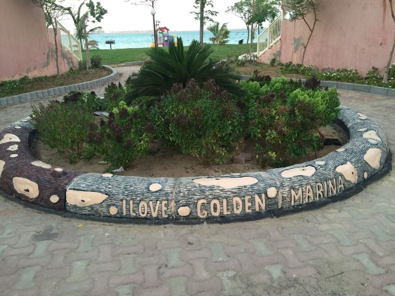 A circular stone pathway with the words "LOVE GOLDEN MARINA" engraved on it, surrounded by greenery and a view of the sea.