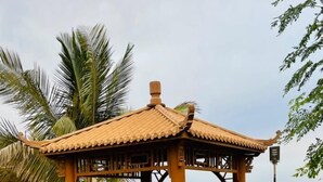 A gazebo with a tiled roof and wooden benches, surrounded by green grass and palm trees, with a view of the sea in the background. - Hotel & Resort Golden Marina Yanbu  فندق ومنتجع جولدن مارينا ينبع إدارة وتشغيل شركة منتجع المرسى الذهبي (Yanbu)