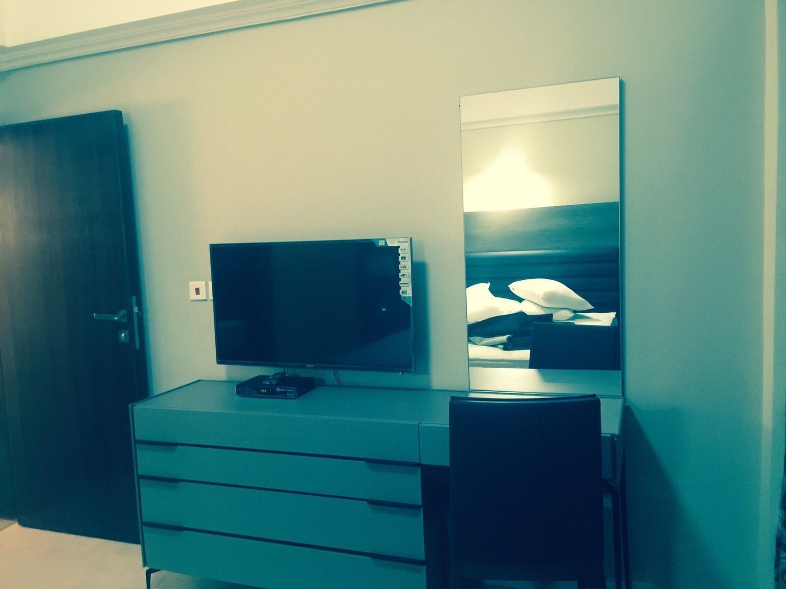 A hotel room with a flat-screen TV on a dresser, a mirror reflecting a bed, and a door to the left.