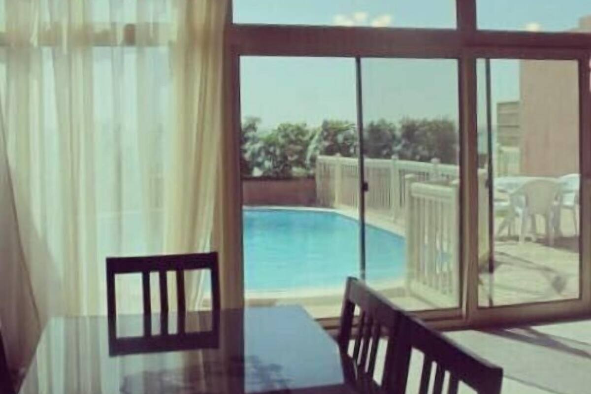 A dining area with a glass table and chairs, overlooking a swimming pool and a fenced outdoor area with chairs.