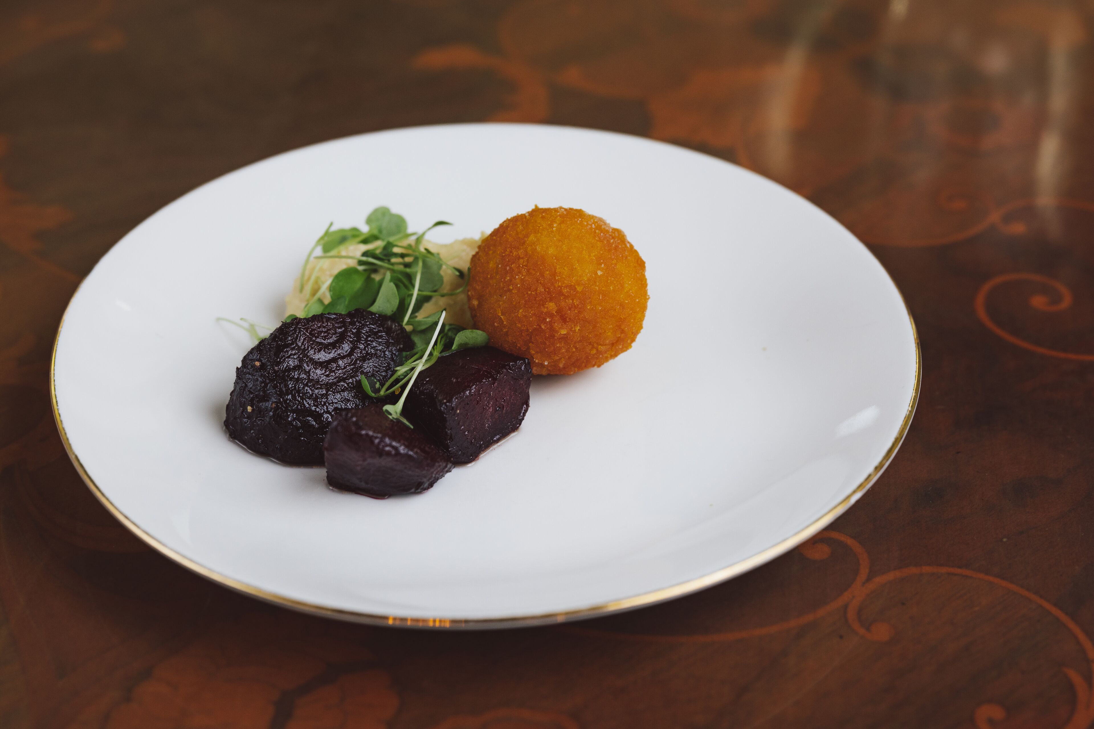 A white plate with a dark meat dish, a golden-brown item, and greens on a wooden table.