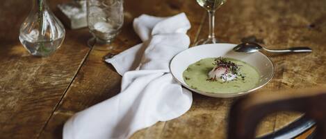 Una mesa de madera con un plato blanco de comida, una copa de vino blanco, un vaso transparente y un florero con flores.