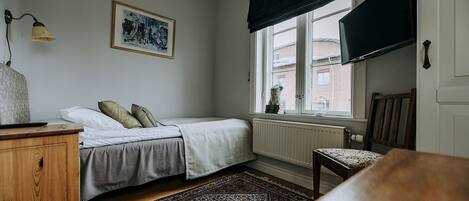 Ein Schlafzimmer mit Bett, Stuhl, Schreibtisch, Heizkörper, einem Fenster mit schwarzem Vorhang und einem Teppich mit Muster. Ein gerahmtes Bild ist an der Wand angebracht.