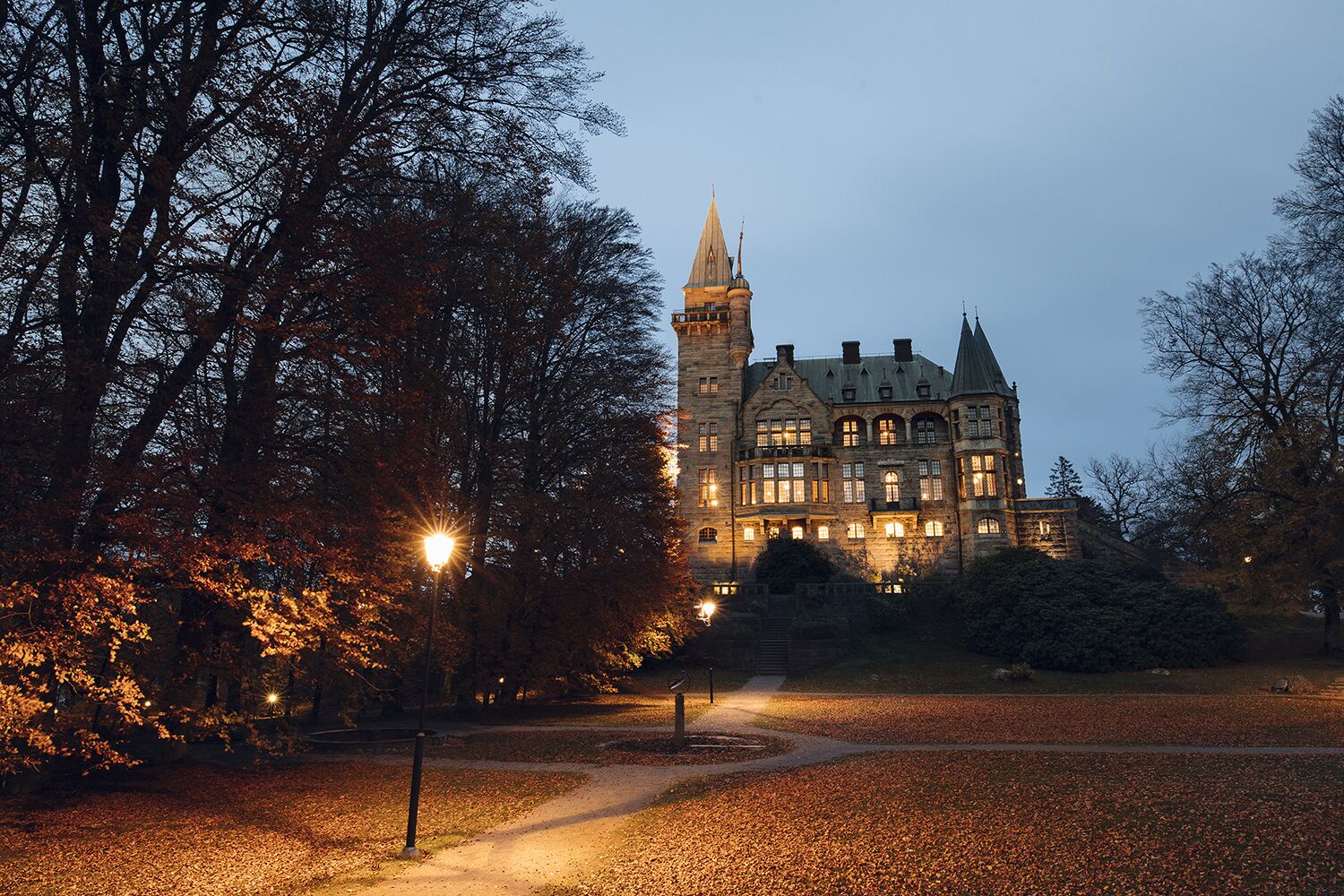 A large, historic building with multiple towers and a prominent central tower, surrounded by trees and a pathway.