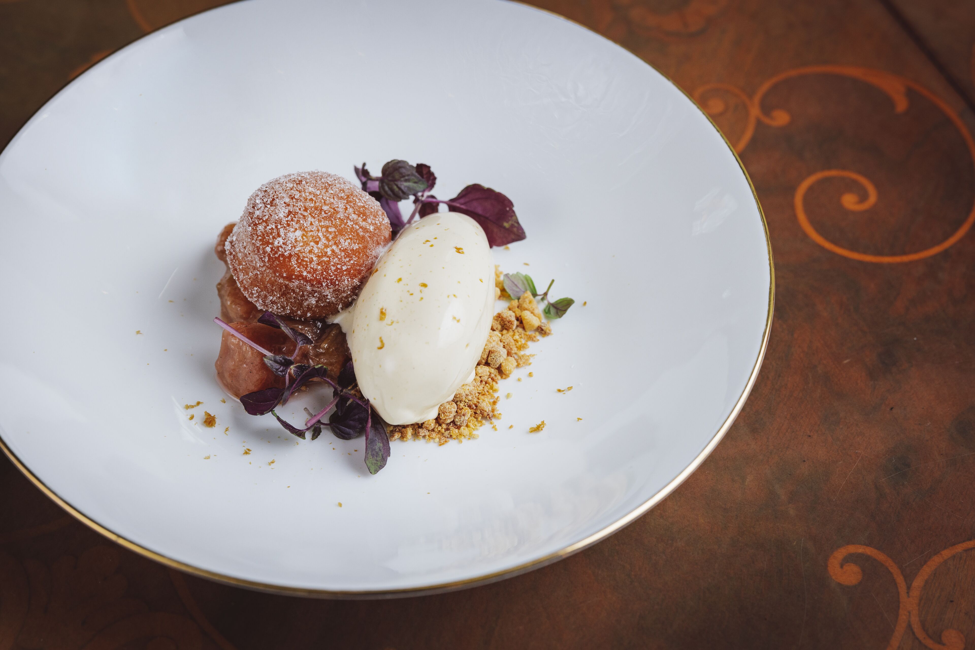 Un plato con un postre que incluye un elemento cubierto de azúcar glas, una bola de helado y un poco de cobertura desmenuzada, posiblemente un crumble o una galleta, sobre un plato blanco con borde dorado.