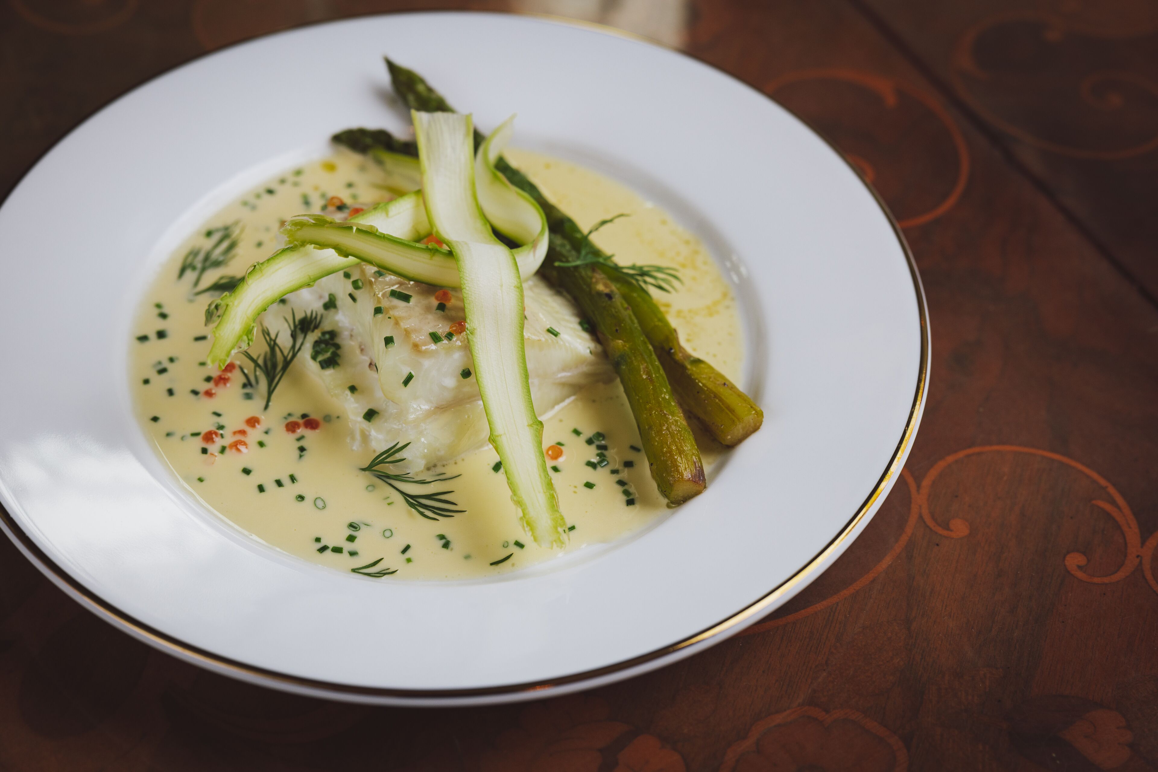 A white plate with a creamy soup, asparagus, and herbs on a wooden table.