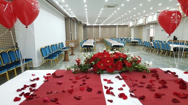 A banquet hall with rows of tables and chairs, a floral centerpiece, and red rose petals on the tables. - The Ancient Mesopotamia Hotel (Batman)