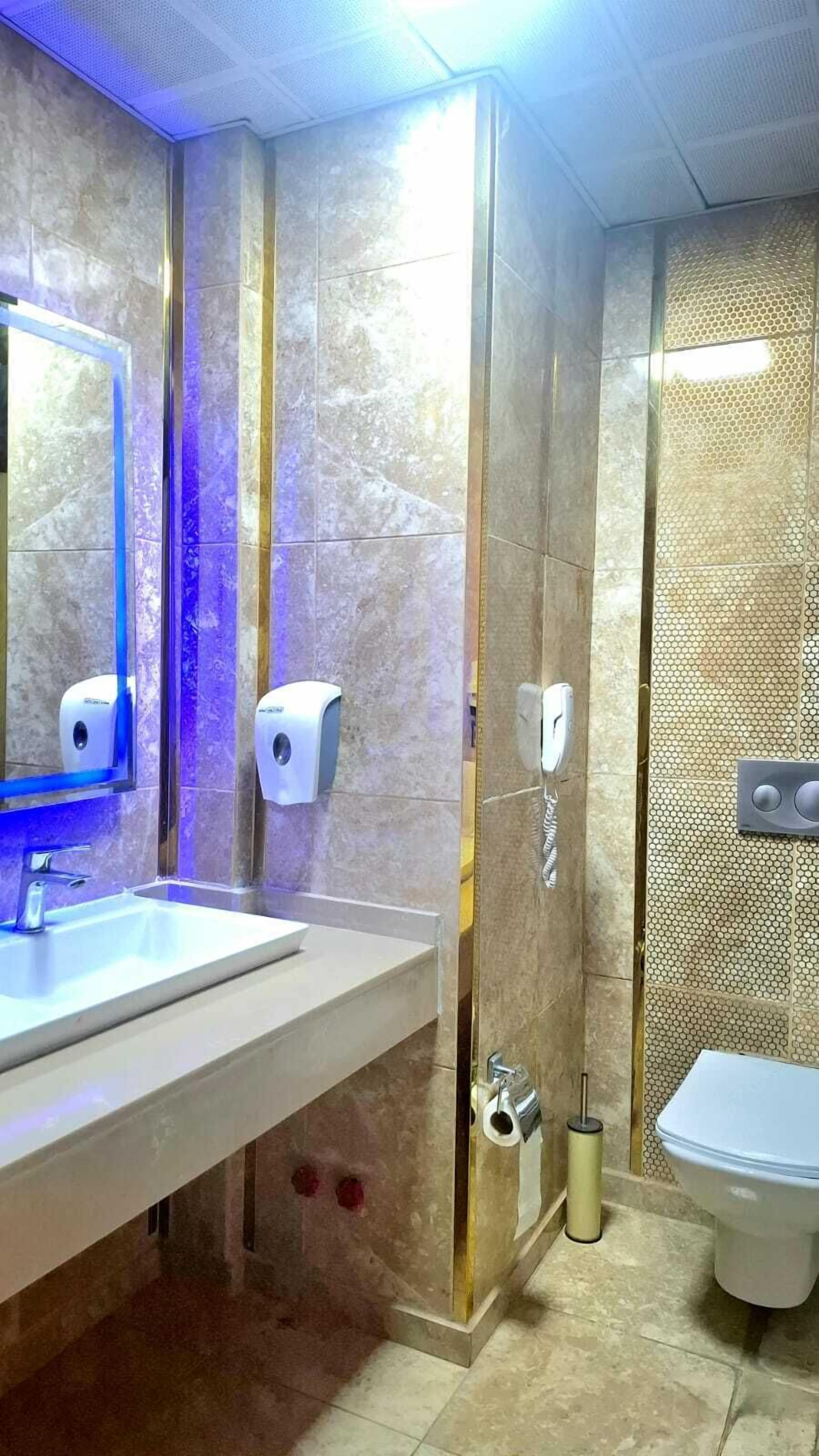Une salle de bain avec un lavabo blanc, deux distributeurs d’essuie-tout, des toilettes et un espace douche doté d’un mur texturé.