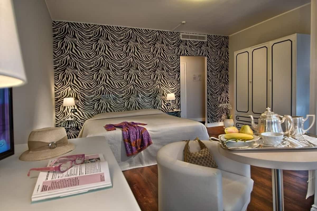 Quarto de hotel com papel de parede listrado em zebra, uma cama, um espaço de estar com uma bandeja de itens de café da manhã e um guarda-roupa.