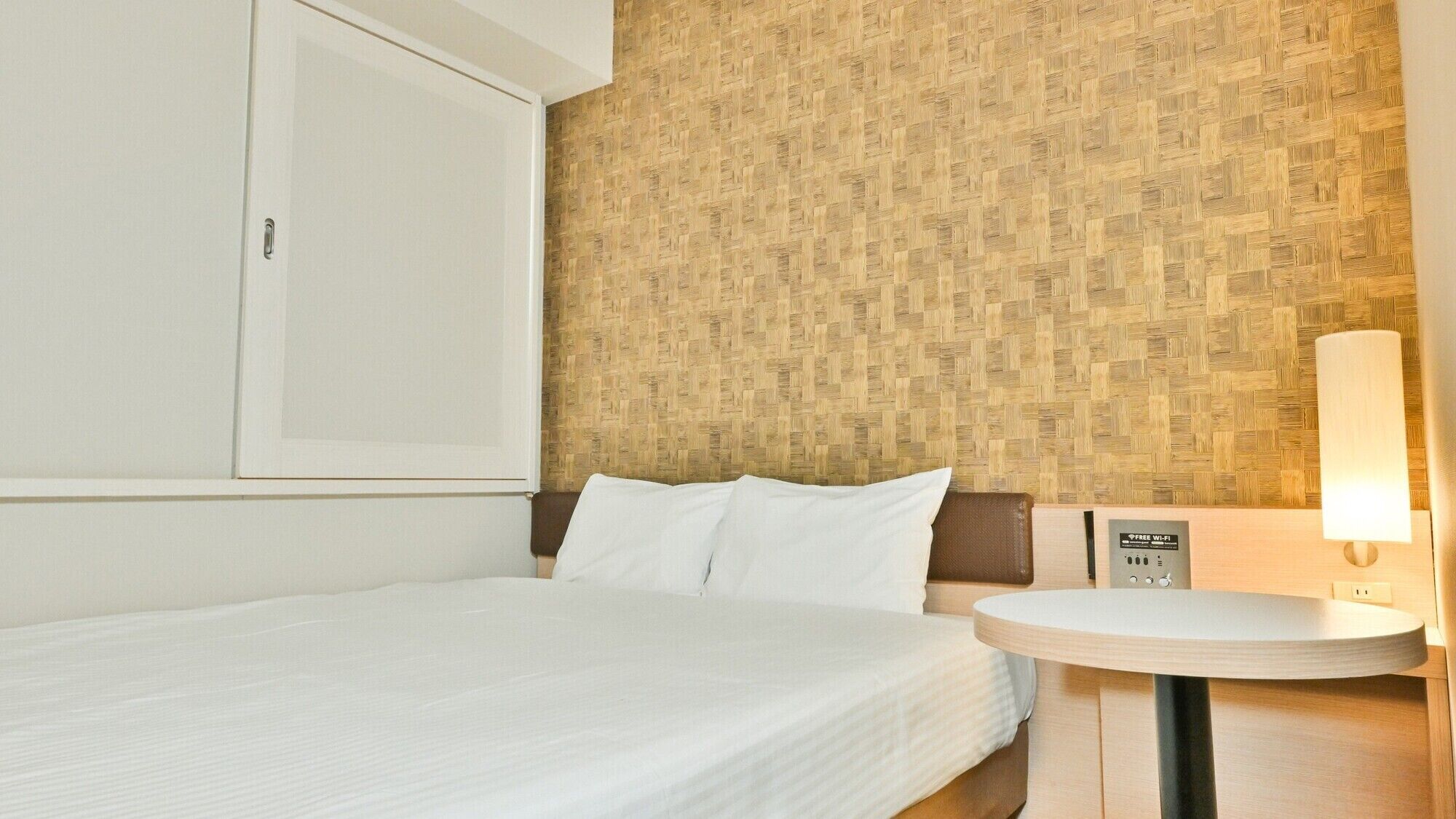 Une chambre d’hôtel avec un lit, une table de chevet avec une lampe, un mur orné d’un motif tissé et une armoire.