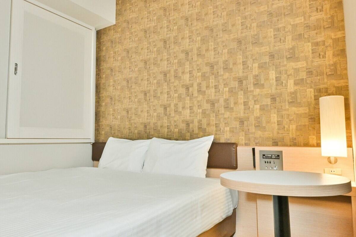 Une chambre d’hôtel avec un lit, une table de chevet avec une lampe, un mur orné d’un motif tissé et une armoire.
