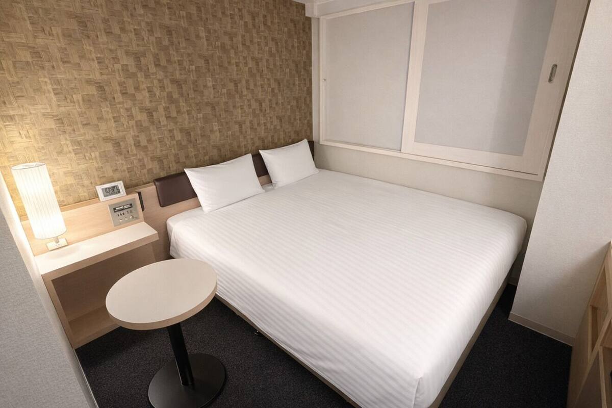 Une chambre d’hôtel avec un lit, une table de chevet avec une lampe, une petite table ronde et une horloge murale.