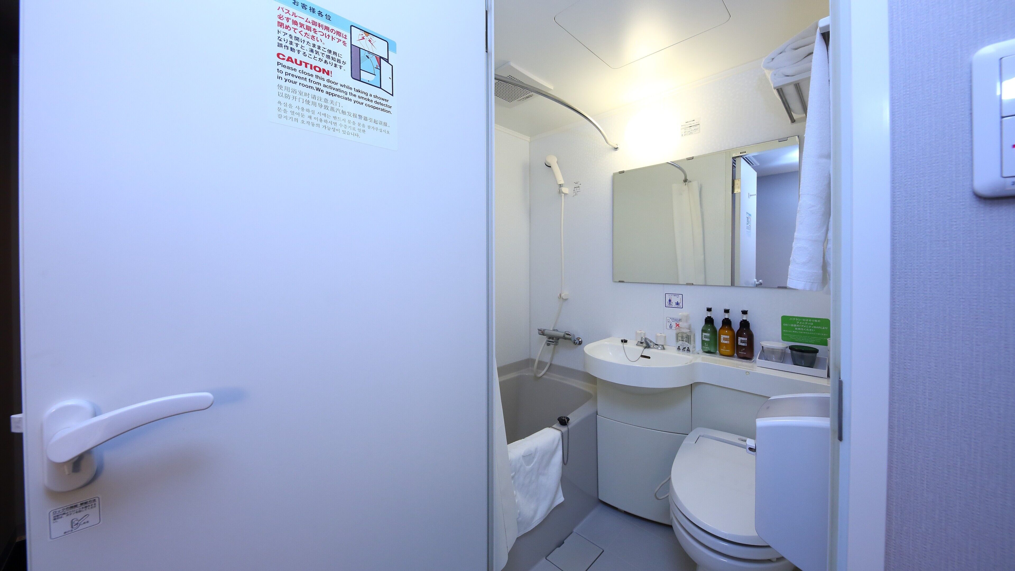 Une petite salle de bain avec des toilettes, un lavabo et une douche. Une serviette est accrochée au mur, et un panneau affiche des instructions.