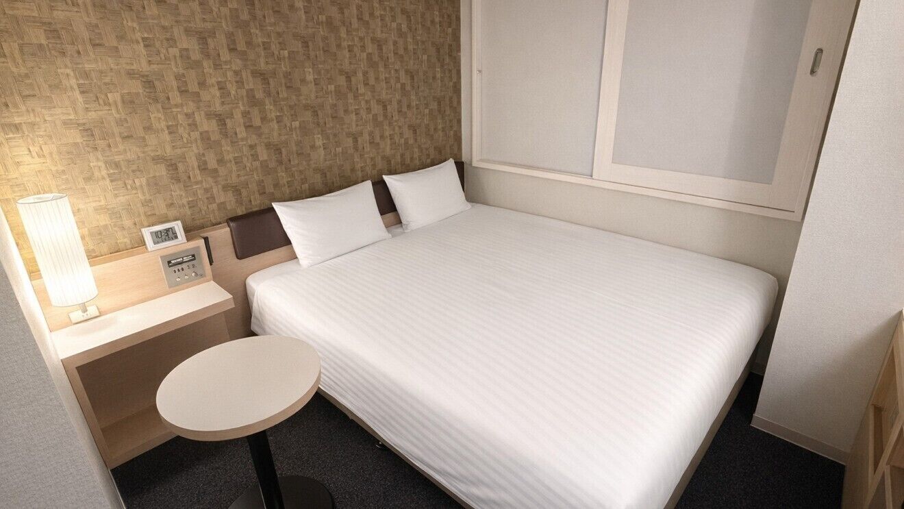 Une chambre d’hôtel avec un lit, une table de chevet avec une lampe, une petite table ronde et une horloge murale.