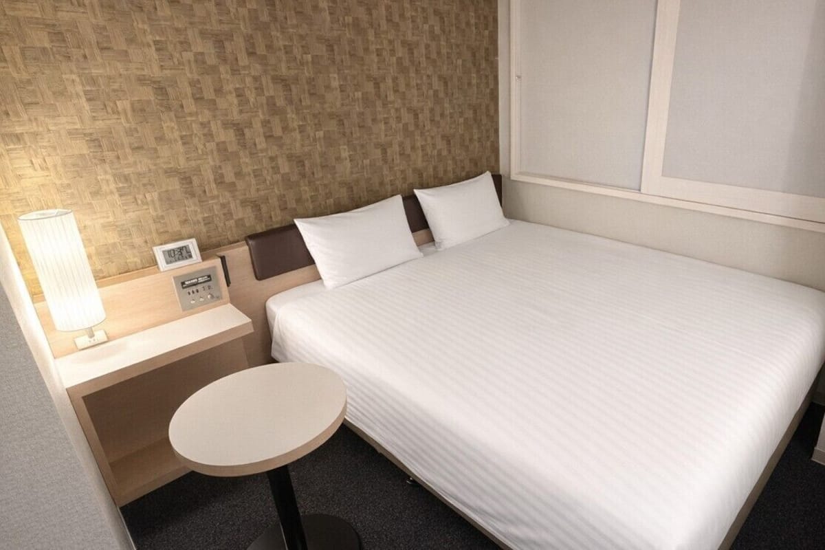 Une chambre d’hôtel avec un lit, une table de chevet avec une lampe, une petite table ronde et une horloge murale.