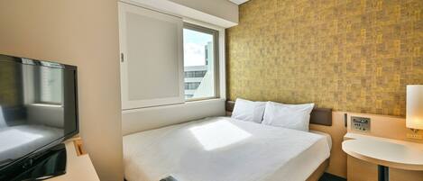 Habitación de hotel con cama, televisión, ventana y un pequeño escritorio con lámpara.
