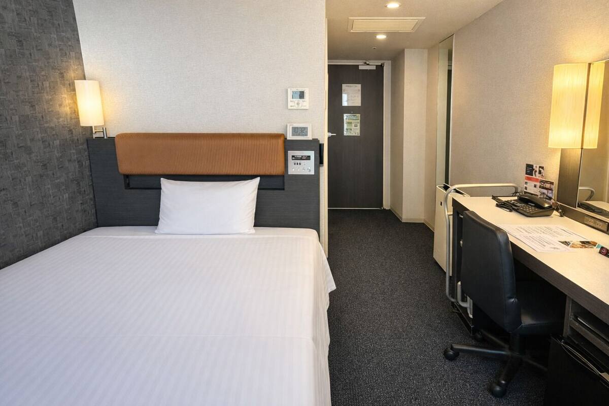 Une chambre d’hôtel avec un lit, un bureau, une chaise, une lampe, une horloge et une porte.