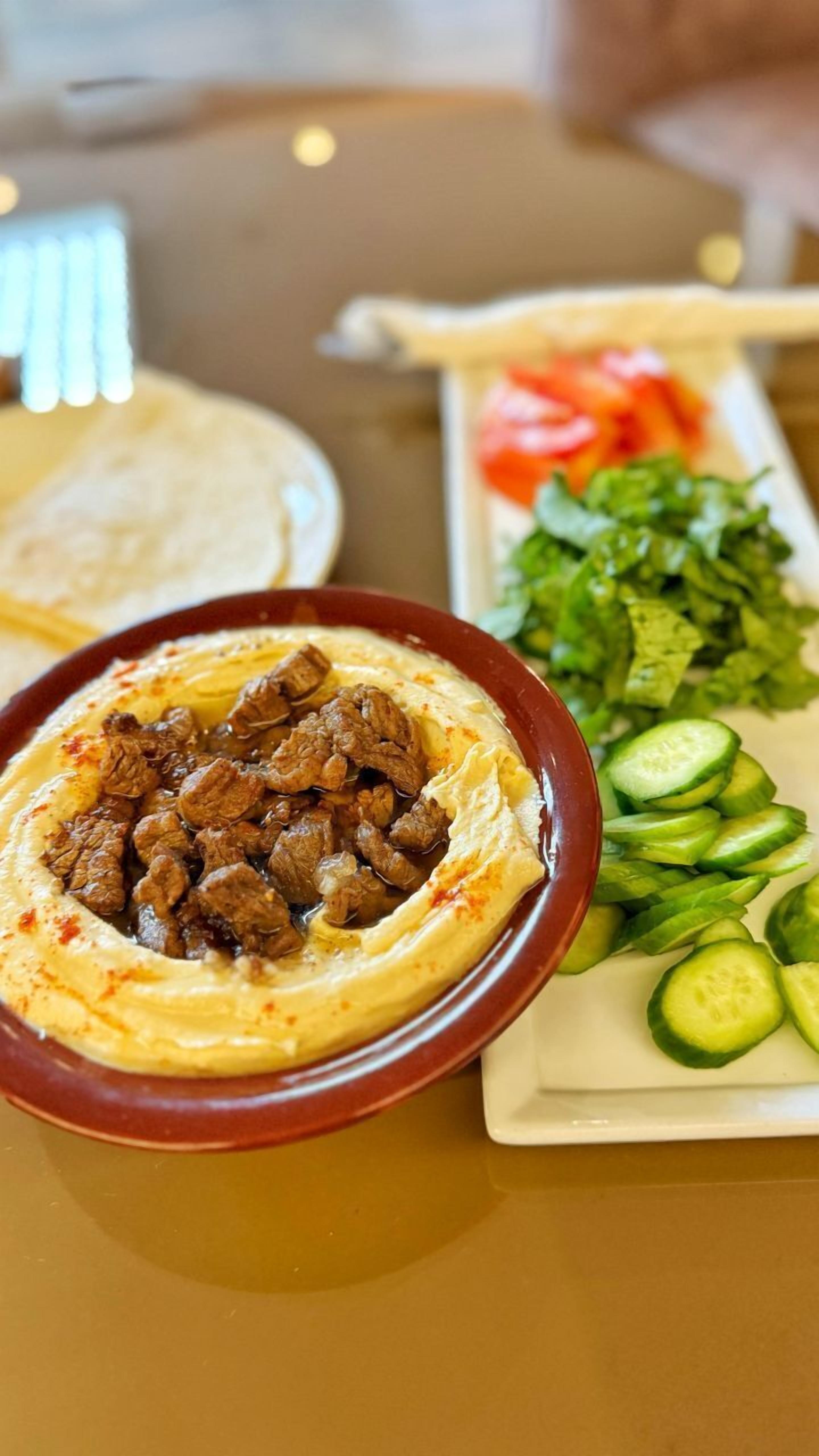 A table with a bowl of hummus topped with meat, a plate of sliced cucumbers, and a plate of vegetables.